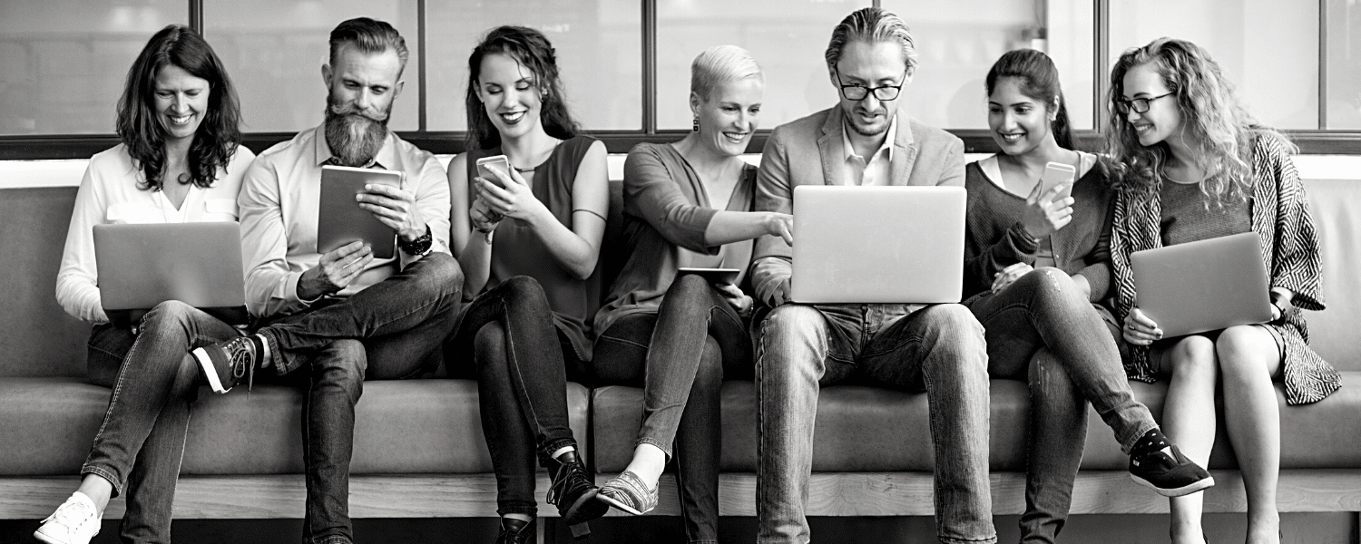 Sieben Personen sitzen in einer Reihe auf einer Couch, lächeln und benutzen verschiedene elektronische Geräte, darunter Laptops, Tablets und Smartphones, was auf eine kollaborative oder technikaffine Gruppenumgebung schließen lässt.